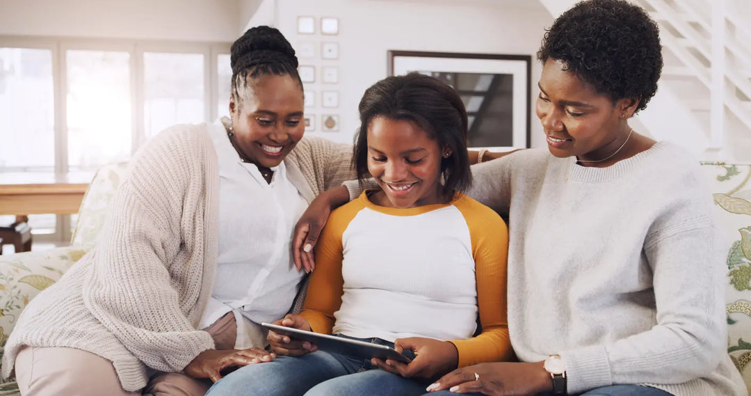 family of black woman gather around teenager with a tablet on the couch