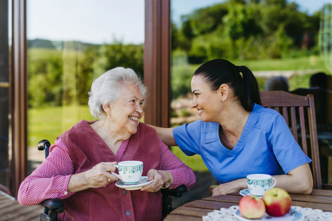 medical professional sits with older woman in a wheelchair outside on a patio drinking tea